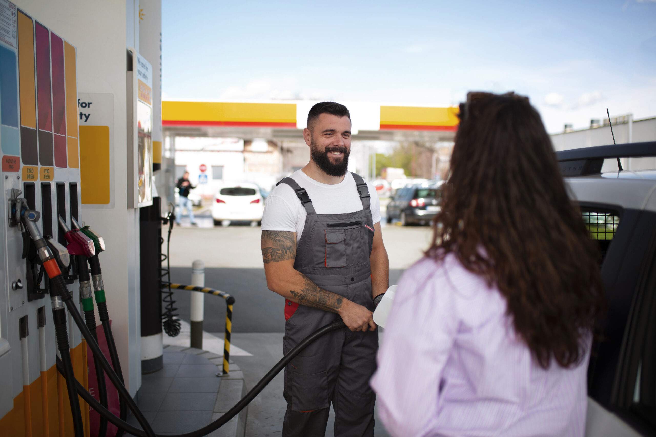 Frentista branco e com barba, usando um macacão cinza, sorri para uma cliente mulher no posto de gasolina