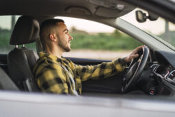 Homem branco, com camisa xadrez amarela com preto, sorrindo ao volante de um carro