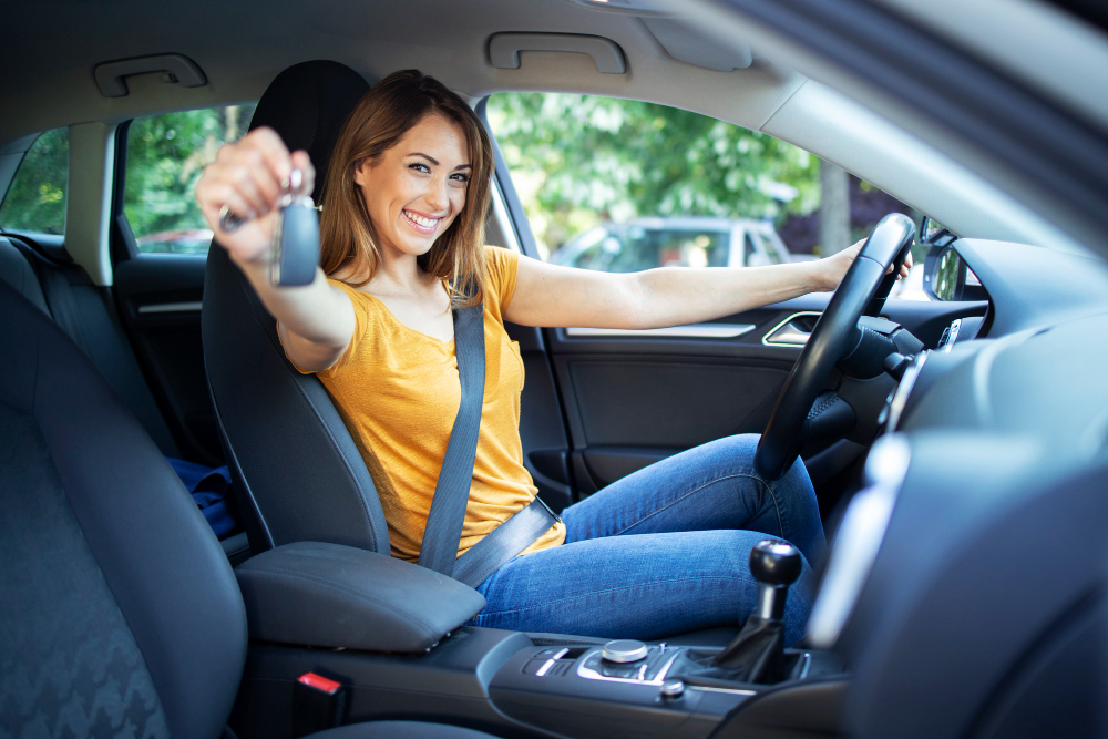 Mulher brranca com camiseta laranja e calça jeans ao volante do carro mostra a chave para a foto